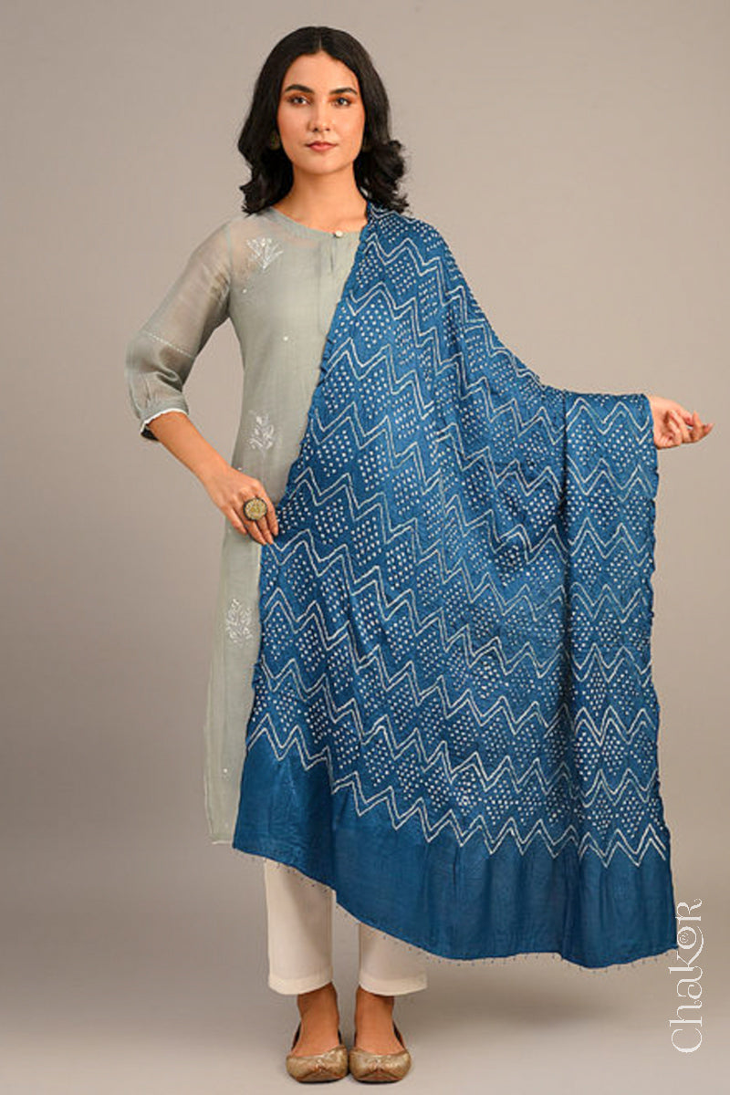 A woman in a grey ethnic outfit, wearing a blue rai bandhej dupatta in gaji silk ,pallu edge detailed with beads, stands gracefully against a light background.Text reads Chakor brand watermark.

