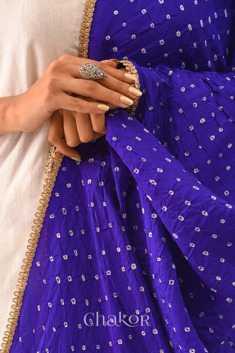 Close-up of a womens hand holding purple Bandhani mulberry silk dupatta with tie-dye dot pattern & detailed with mirror work & golden lace on edge ,draped over a white kurta.