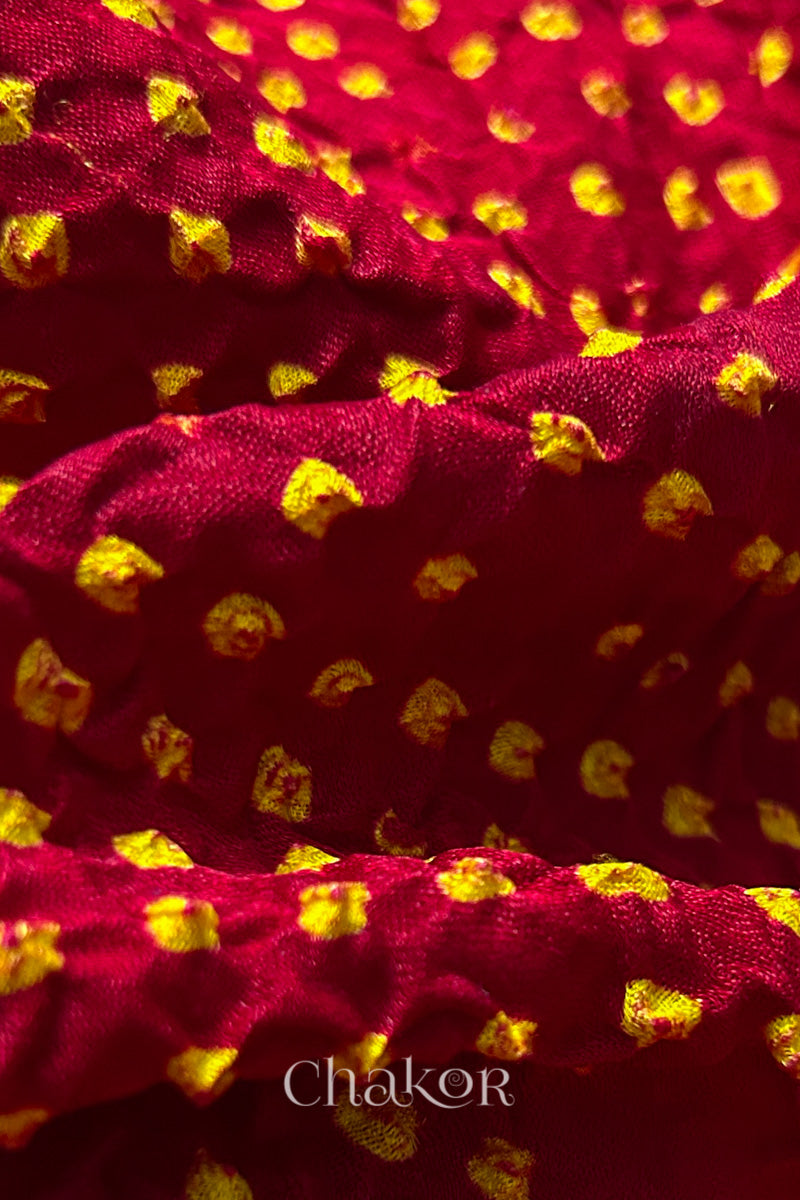 Close-up of a rai bandhej dupatta in red featuring  yellow bandhani dots.Text reads Chakor brand watermark.
