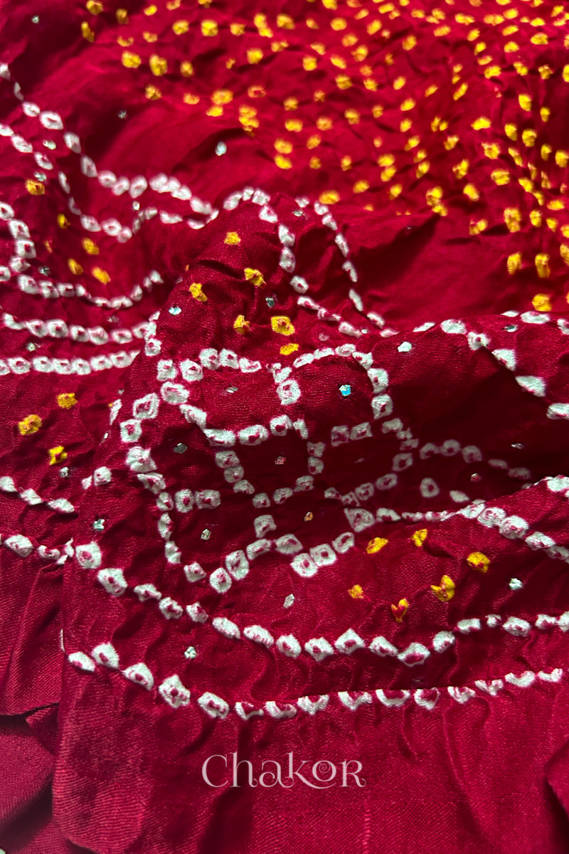 Close-up of a rai bandhej dupatta in red featuring white & yellow bandhani dots along with mukaish embroidery.Text reads Chakor brand watermark.
