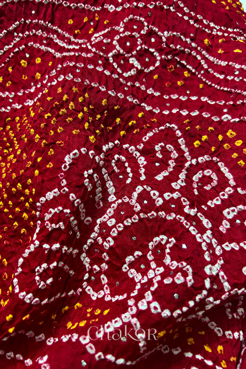Close-up of a rai bandhej dupatta  in red showing the peacock pattern featuring white & yellow bandhani dots along with mukaish embroidery.Text reads Chakor brand watermark.
