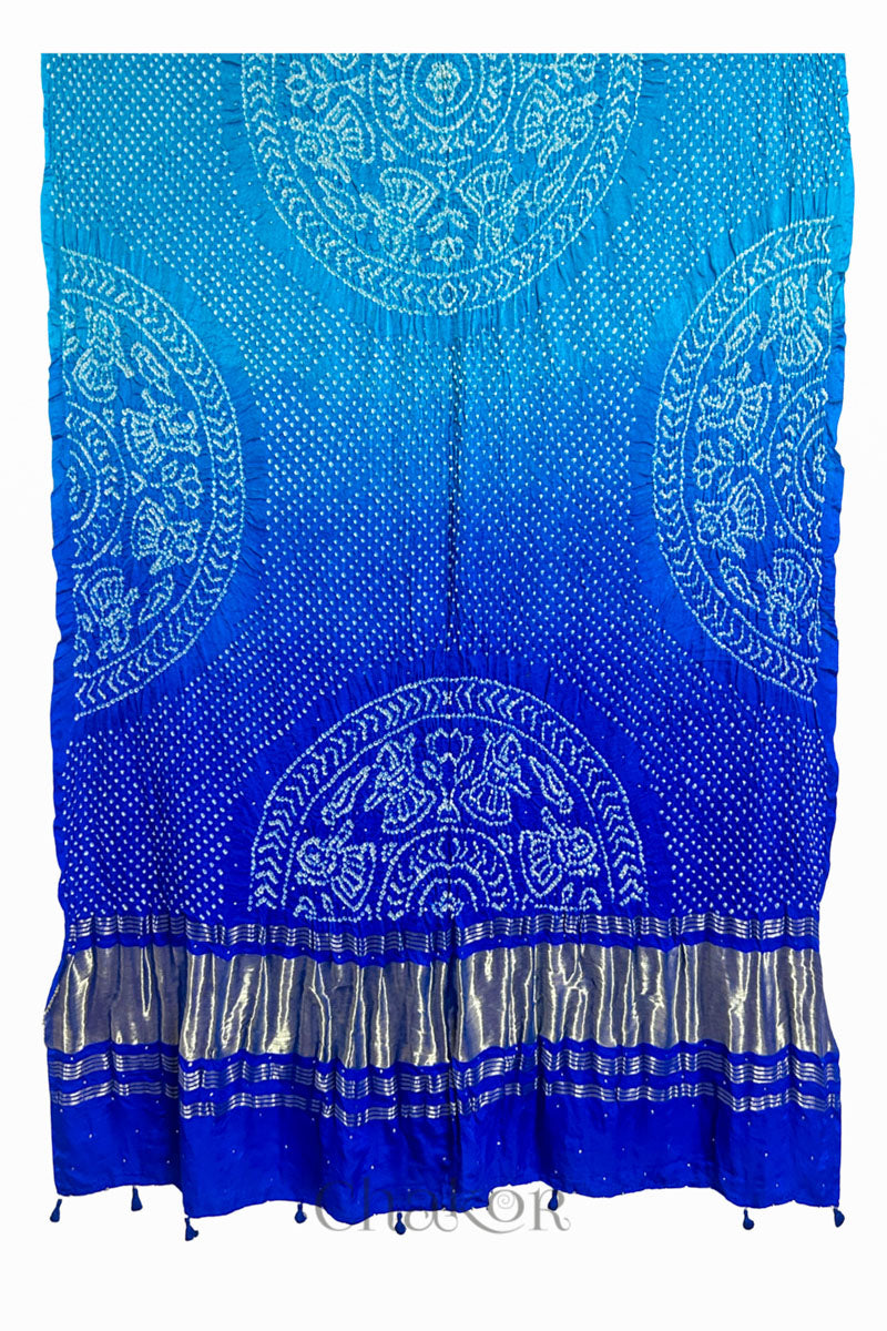 A vibrant blue ombre dyed dupatta showcasing intricate rai bandhej with a shimmering gold band on the pallu and tassels on the hem. Text reads Chakor brand watermark.
