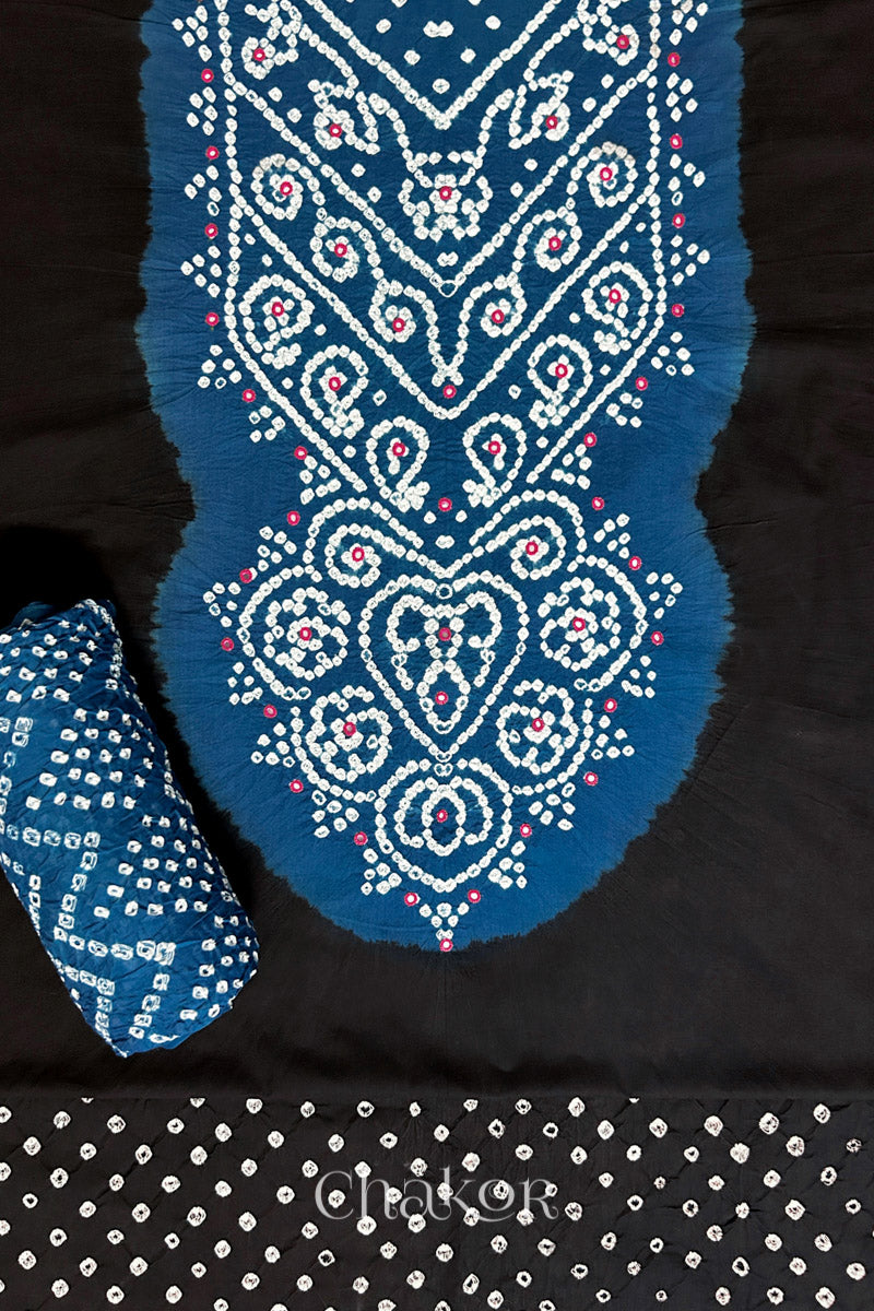 A classic Black set fabric featuring mirror embroidered contrast yoke in indigo blue, matching coloured bandhani bottom and indigo blue dupatta rolled beautifully and placed on the fabric. Text reads Chakor.
