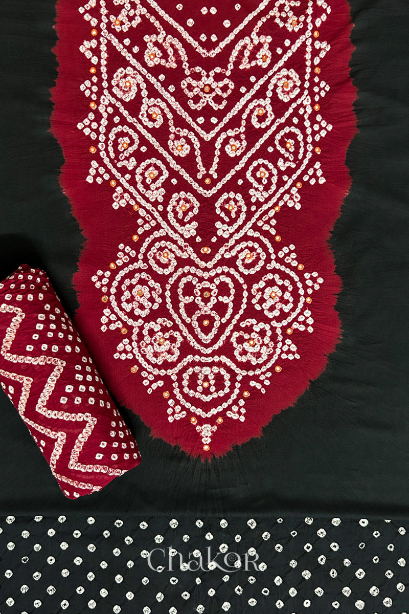 A classic Black set fabric featuring mirror embroidered contrast yoke in red, matching coloured bandhani bottom and red dupatta rolled beautifully and placed on the fabric. Text reads Chakor.