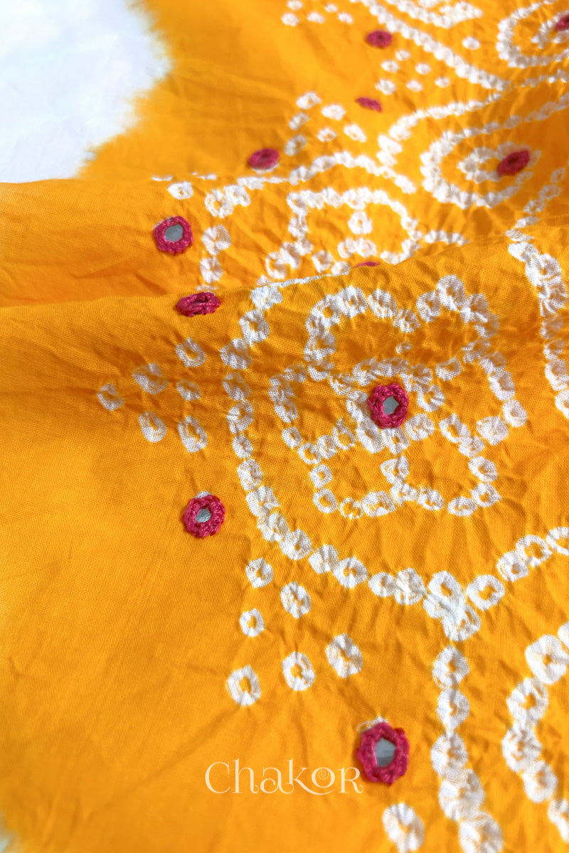 Close-up of yellow-white fabric adorned with intricate traditional bandhani patterns and pink mirror embroidery, showcasing beautiful craftsmanship.