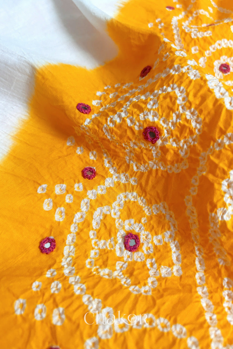 Close-up of yellow-white fabric adorned with intricate traditional bandhani patterns and pink mirror embroidery, showcasing beautiful craftsmanship.