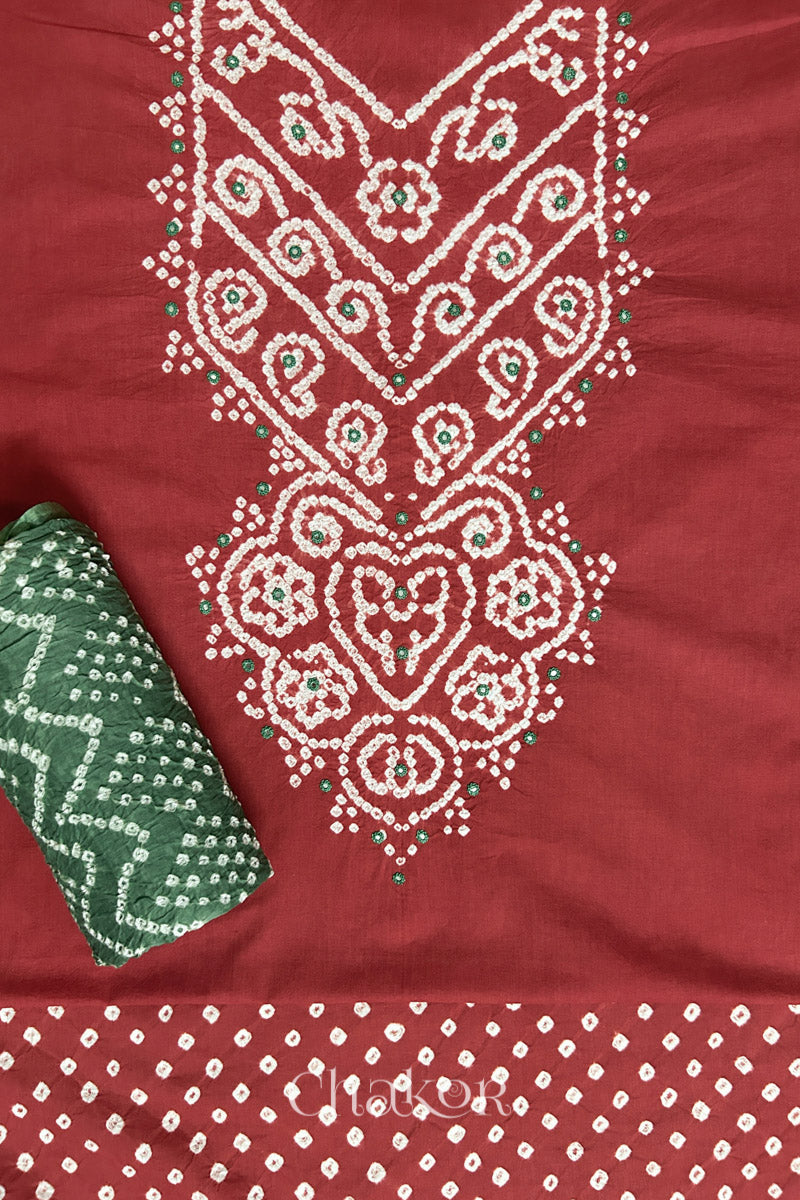 A stylish brick red set fabric featuring mirror embroidered yoke, bandhani bottom and contrast moss green dupatta rolled beautifully and placed on the fabric. Text reads Chakor.