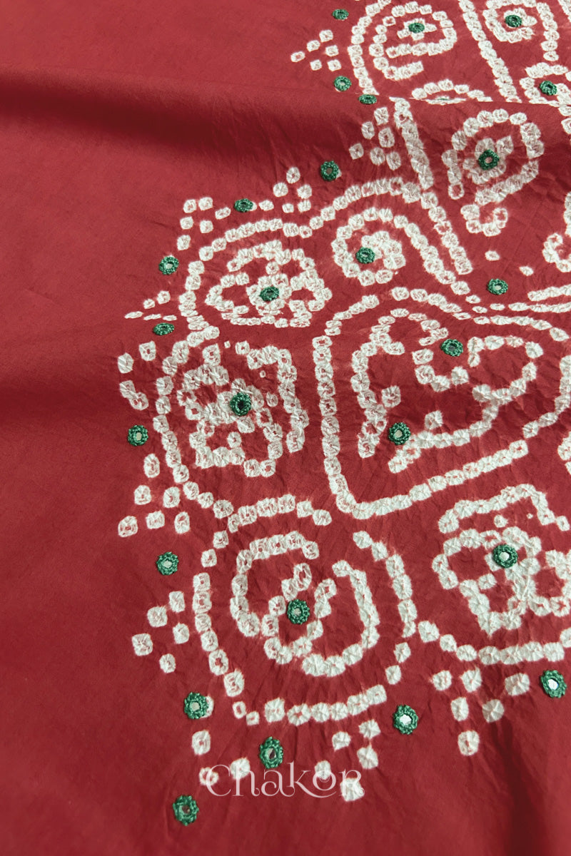 Close-up of  brick red fabric adorned with intricate traditional bandhani patterns and green mirror embroidery, showcasing beautiful craftsmanship.