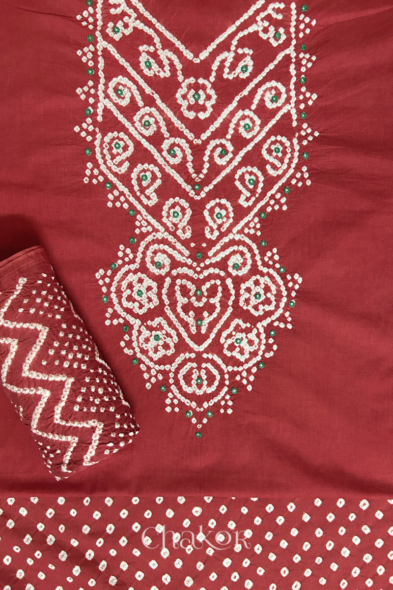 A stylish brick red set fabric featuring mirror embroidered yoke, bandhani bottom and dupatta rolled beautifully and placed on the fabric. Text reads Chakor.