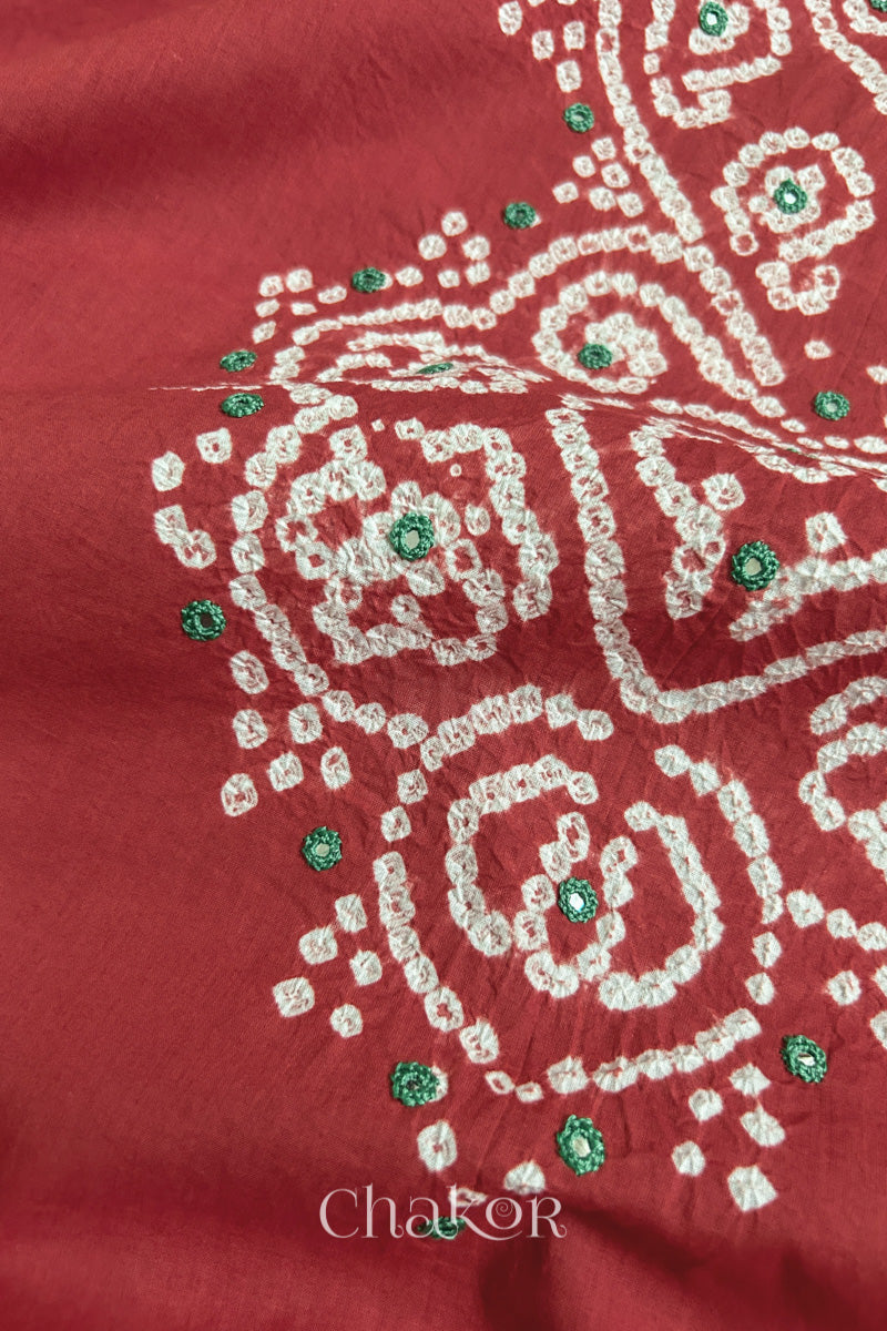 Close-up of  brick red fabric adorned with intricate traditional bandhani patterns and green mirror embroidery, showcasing beautiful craftsmanship.
