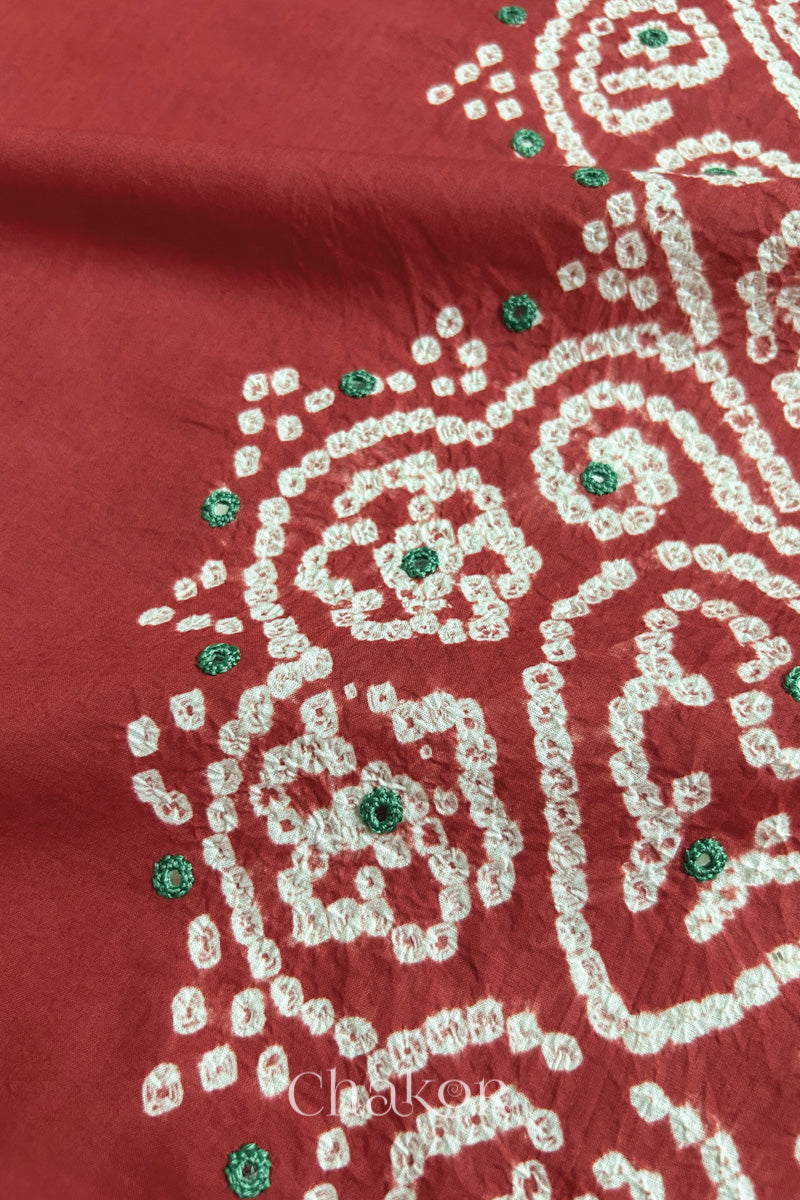 Close-up of  brick red fabric adorned with intricate traditional bandhani patterns and green mirror embroidery, showcasing beautiful craftsmanship.