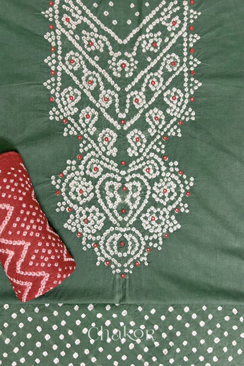 A stylish moss green set fabric featuring mirror embroidered yoke, bandhani bottom and contrast brick red dupatta rolled beautifully and placed on the fabric. Text reads Chakor.