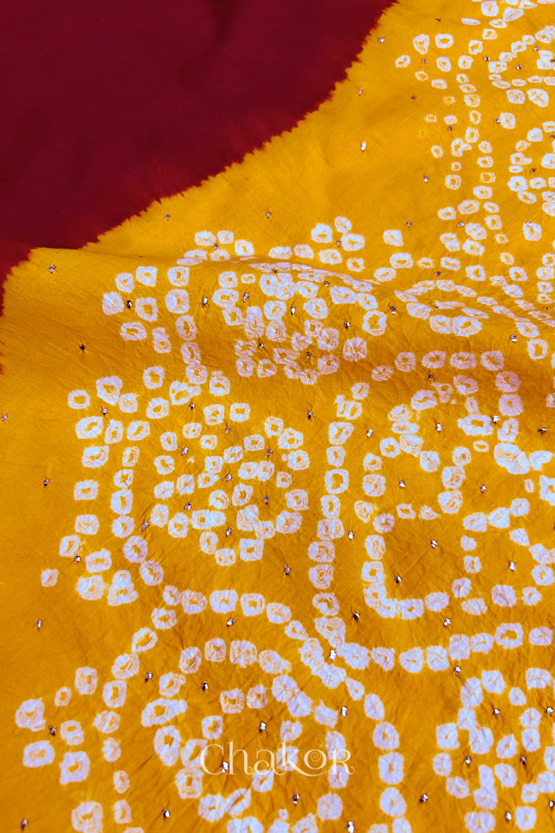 Close-up of red fabric adorned with intricate traditional bandhani patterns and sparkly mukaish embroidery on yellow yoke, showcasing beautiful craftsmanship.
