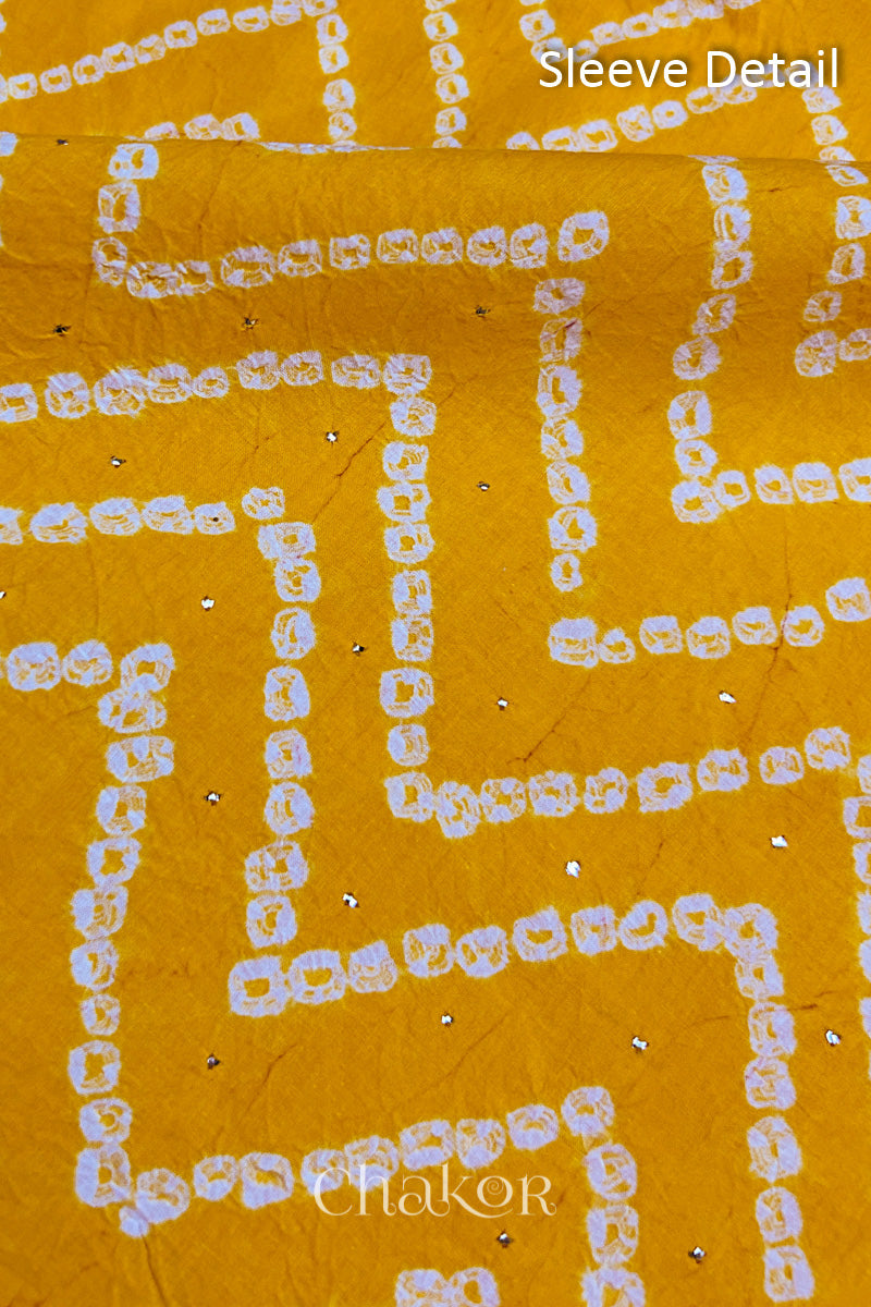Close-up of yellow bandhani fabric featuring chevron tie-dye design, showcasing sparkly mukaish embroidery with Chakor brand watermark. Text reads sleeve details.
