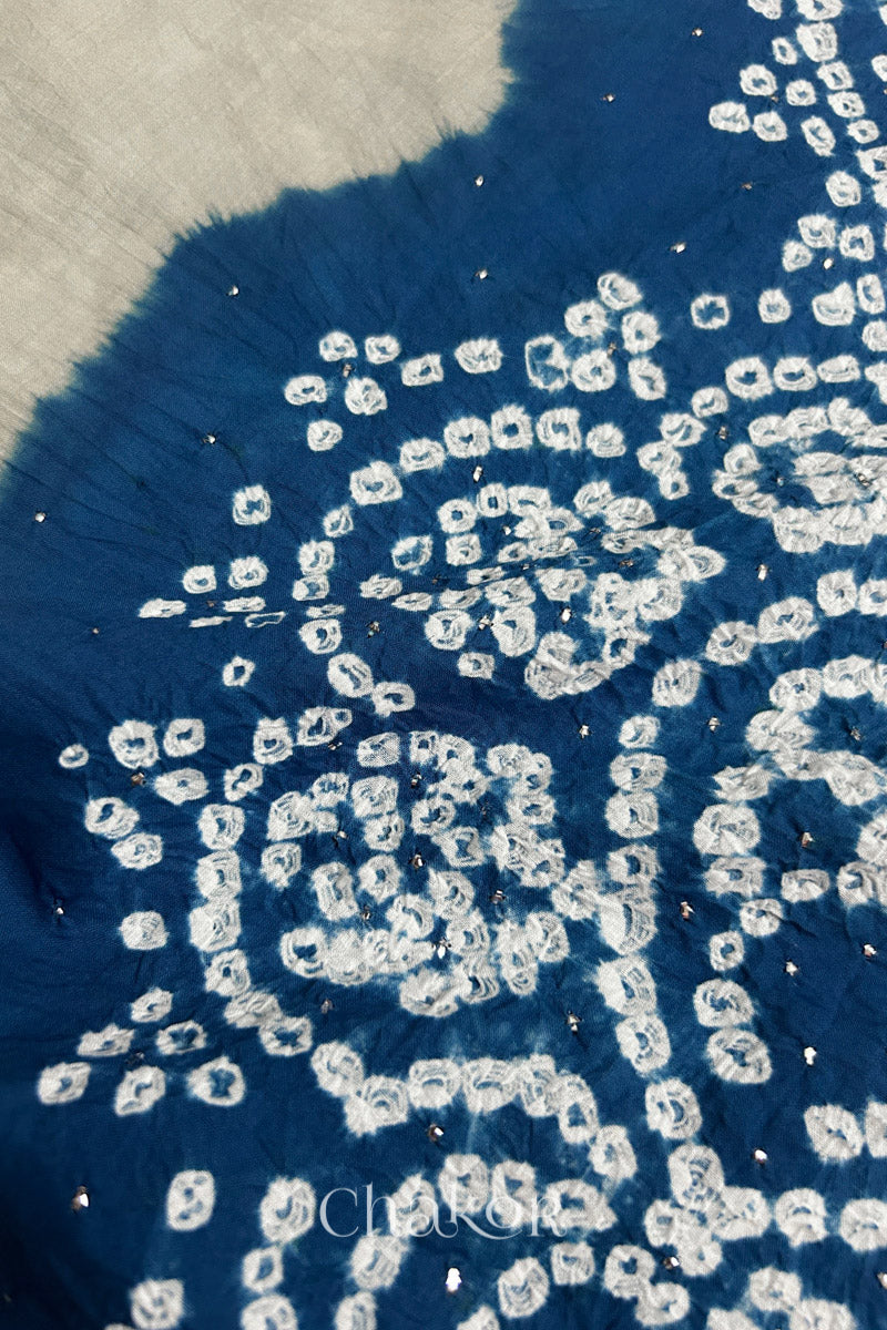 Close-up of  light grey fabric adorned with intricate traditional bandhani patterns and sparkly mukaish embroidery on indigo blue yoke, showcasing beautiful craftsmanship.
