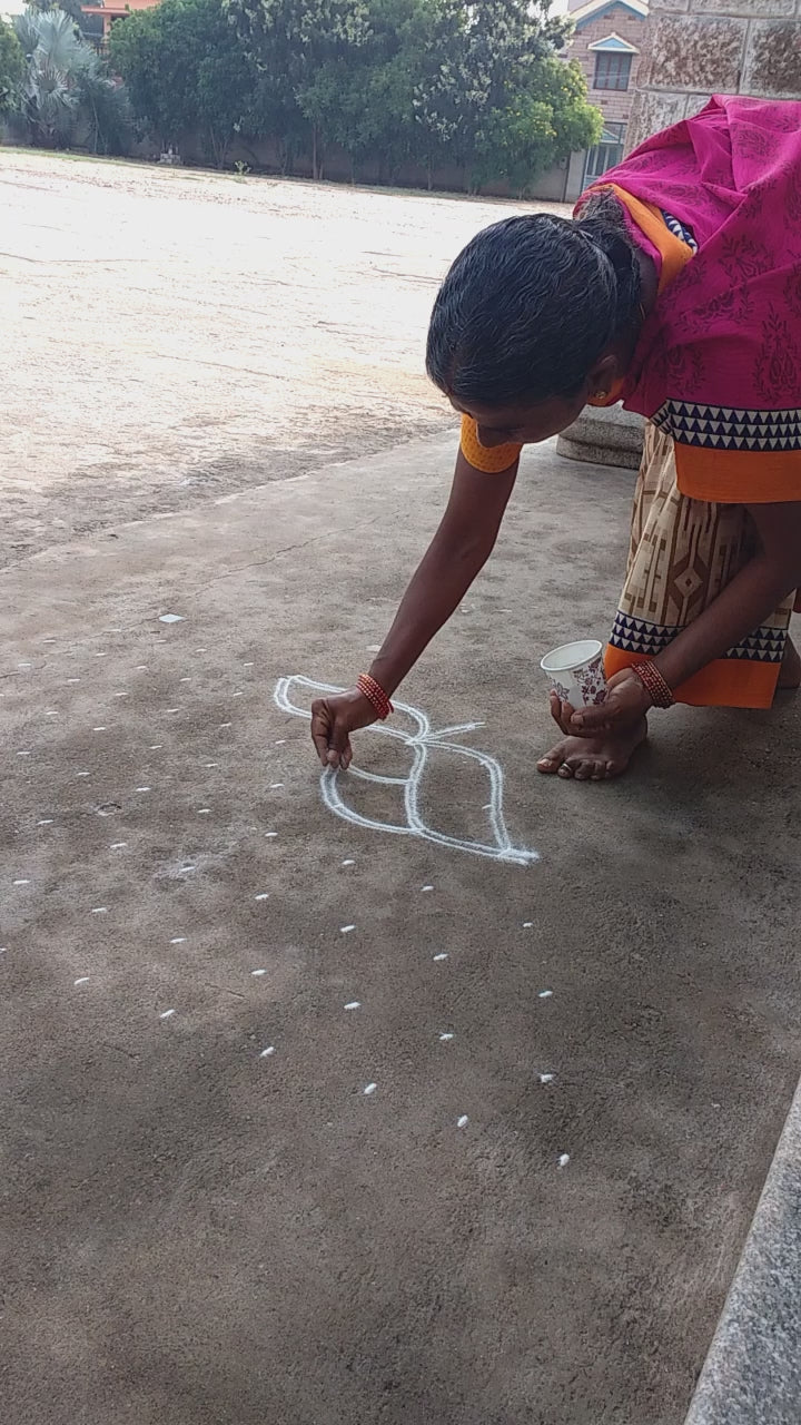 A woman in a simple saree crouches at the entrance of a craft exhibit, gently arranging a rangoli design with care. Her quiet gestures speak of warmth and intention, captured in a soft, reflective tribute to unseen acts of kindness.